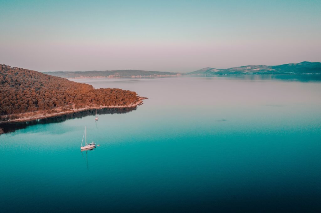 Tranquil aerial view of the Croatian coastline with a lone sailboat on calm blue waters, surrounded by lush green hills.